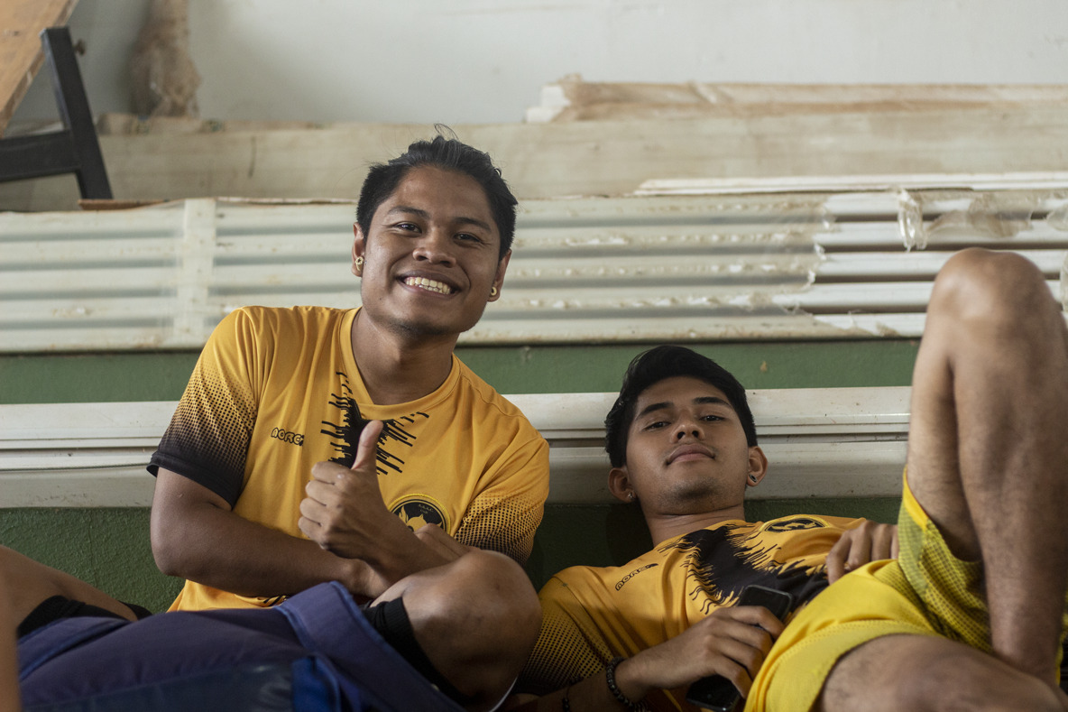 dois homens com camiseta amarela. o homem a esquerda sorri e faz um joinha com a mão, o homem a esquerda está semideitado.