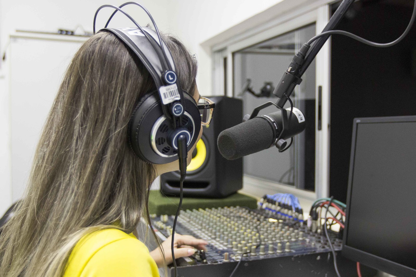 Mulher de costas, cabelo liso, com fone de ouvido estilo headphone, em frente a um microfone  condensador e um computador. Ao seu lado esquerdo está uma mesa de som e um caixa de som.