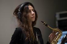Em Plano Médio, vista lateral da multi-instrumentista PJ Harvey com fones-de-ouvido segurando um saxofone.