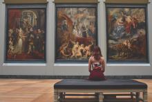 foto de mulher sentada em banco de museu admirando três grandes quadros a sua frente