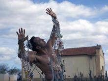 Na imagem, o ator Irandhir Santos aparece dançando ao ar livre, com os braços voltados para o alto, em uma das cenas do filme "A história da Eternidade". Ao fundo, vê-se o céu e o muro de uma igreja.