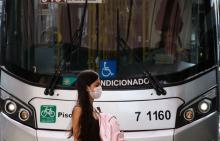 Na imagem, vê-se uma mulher de máscara, logo atrás, a parte da frente de um ônibus
