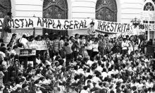 Na imagem, que está em preto e branco, uma multidão reunida na Cinelândia, Rio de Janeiro, parte do grupo está sentado em uma escadaria e parte está em pé. Acima, uma faixa onde lê-se "Anistia ampla, geral e irrestrita".