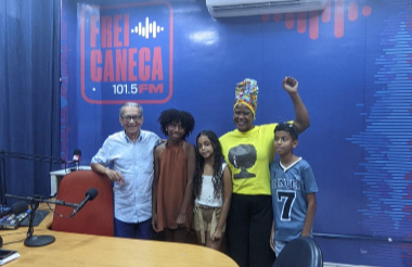 Cinco pessoas posam lado a lado no estúdio da Frei Caneca FM. Ao fundo, há uma parede azul com o logotipo da rádio. Uma mulher negra, de blusa amarela e turbante colorido, levanta o punho direito. À frente, aparecem uma mesa de madeira, microfones e cadeiras.